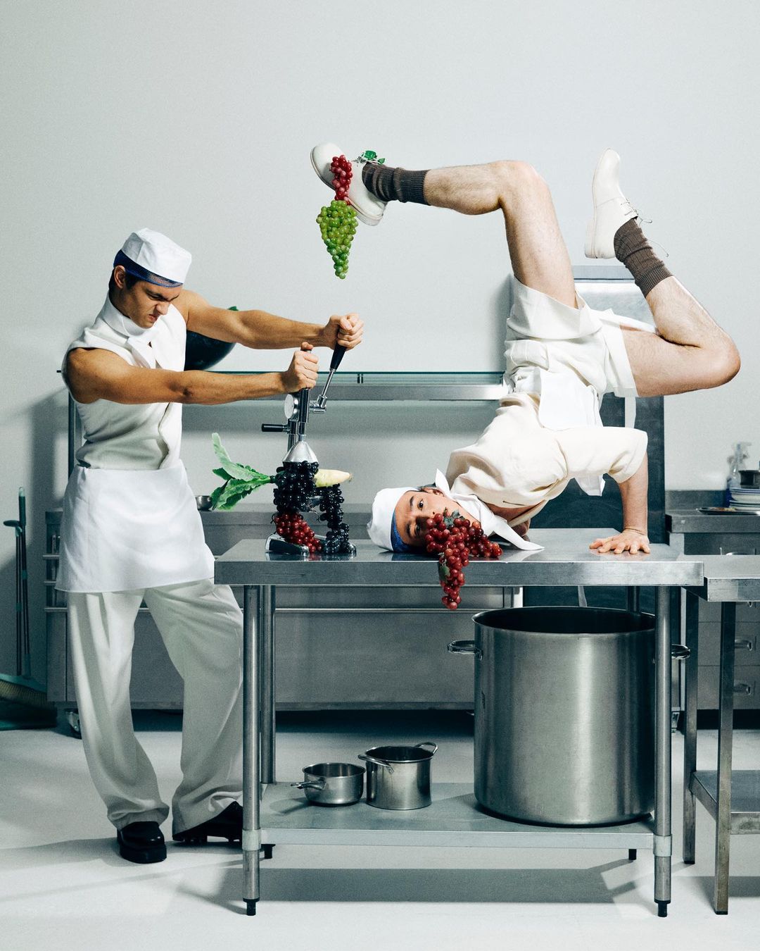 Two adult males in chef whites striking comical poses on stainless steel kitchen worktops with kitchenware and green and red grapes.