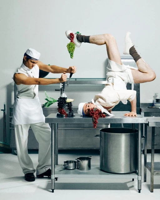 Two adult males in chef whites striking comical poses on stainless steel kitchen worktops with kitchenware and green and red grapes.