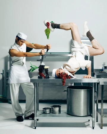 Two adult males in chef whites striking comical poses on stainless steel kitchen worktops with kitchenware and green and red grapes.