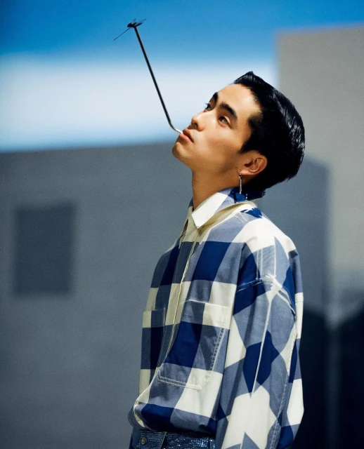 Adult male poses indoors with a straw in his mouth, wearing a blue and white checkered shirt. The background appears to be architectural.