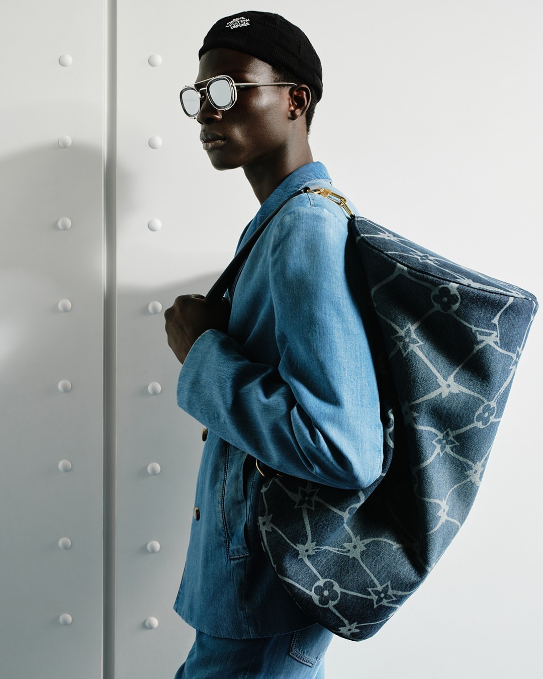 Dark-skinned male model poses in a blue denim jacket and jeans, carrying a blue and gray patterned shoulder bag indoors.