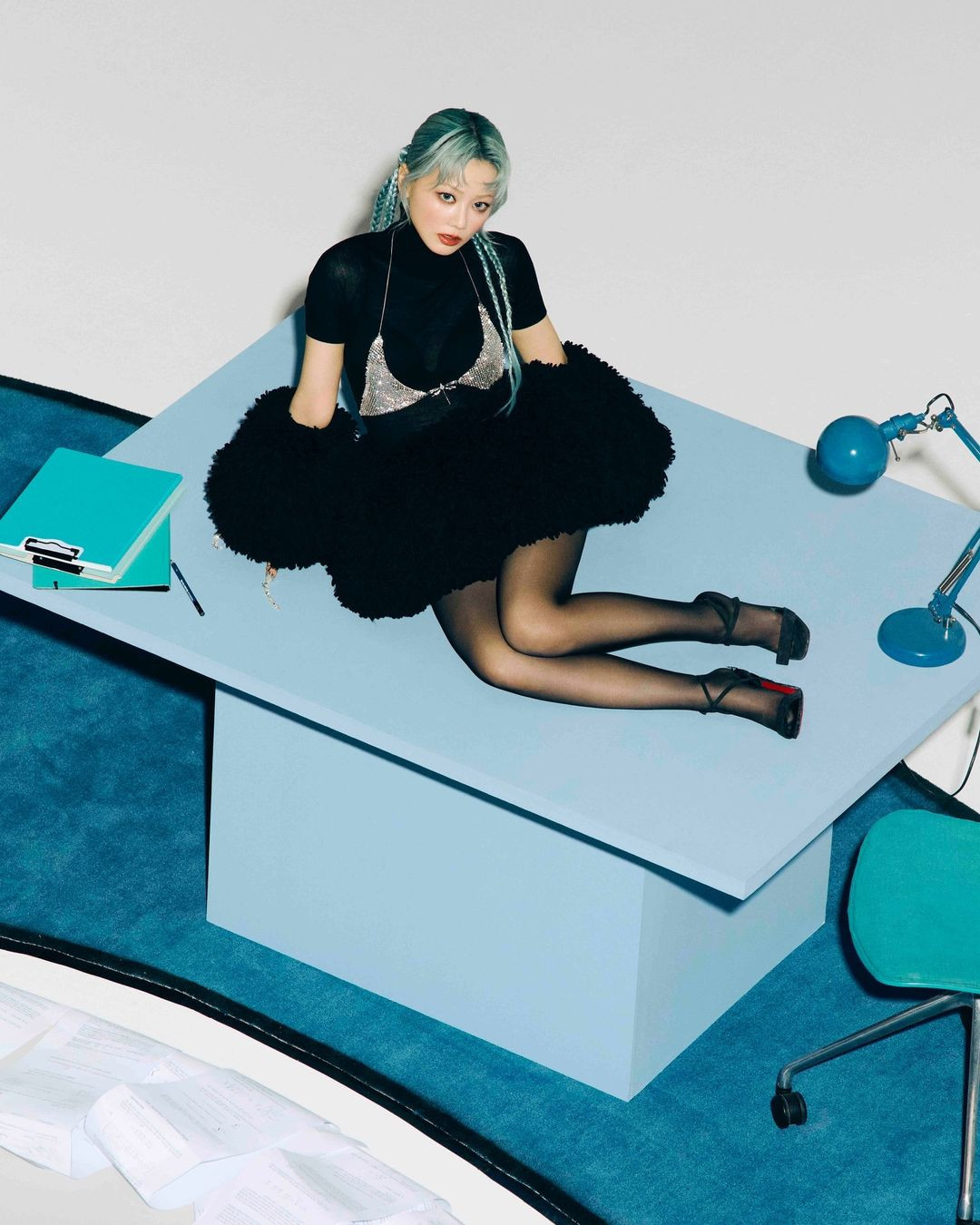 Woman with mint hair poses sitting on a blue desk with a turquoise clipboard and lamp, wearing a black top and beaded bralette.