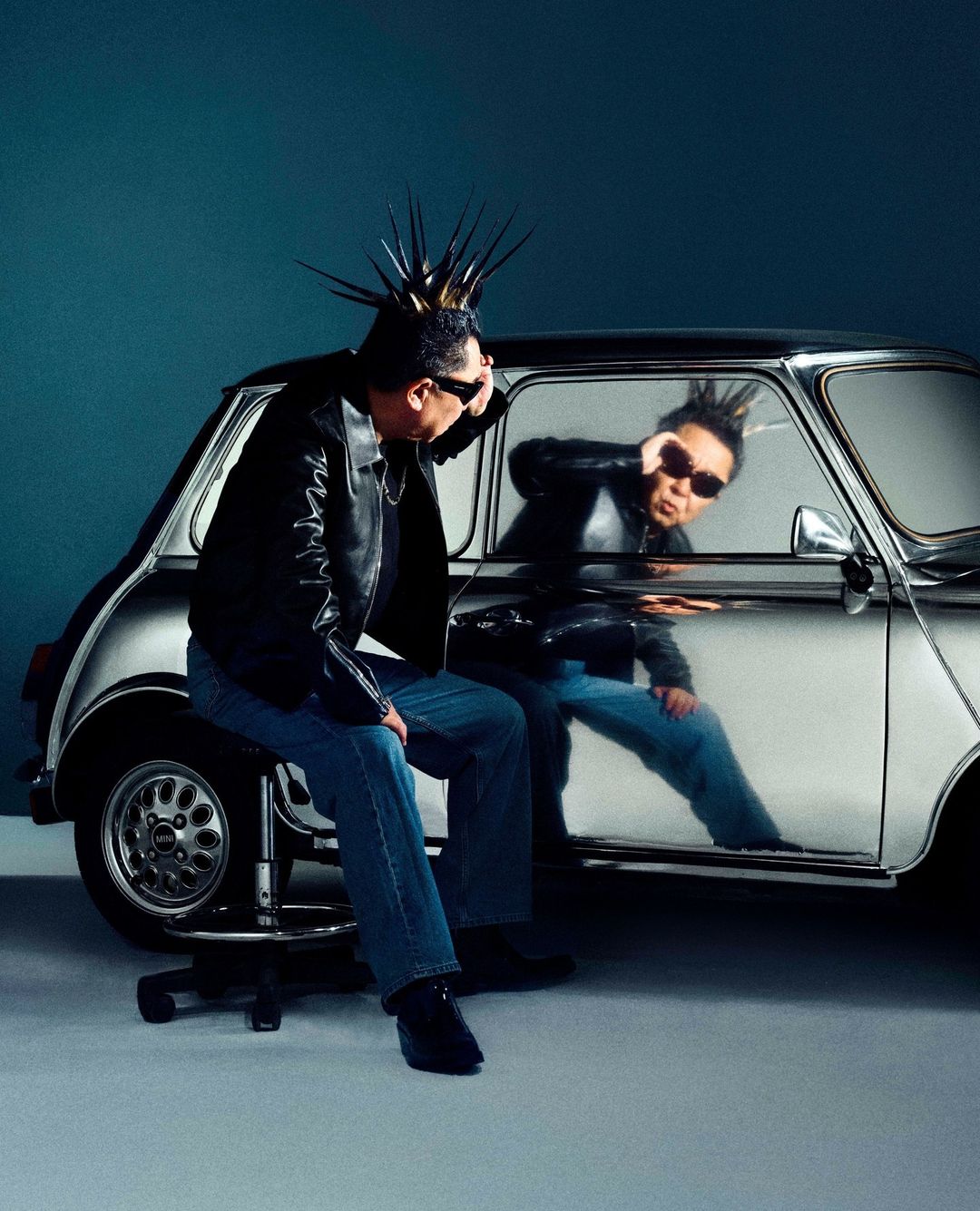 Adult with a mohawk sits next to a reflective Mini Cooper in a studio, wearing sunglasses, a leather jacket, and jeans, creating a retro vibe.