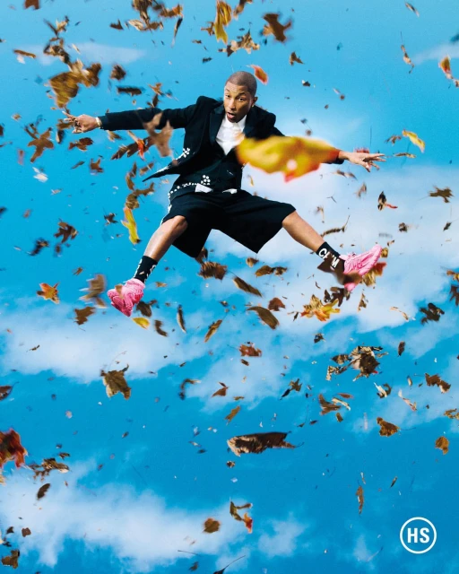 A man in a black suit jacket and shorts jumps in the air amidst falling autumn leaves against a blue sky with clouds.