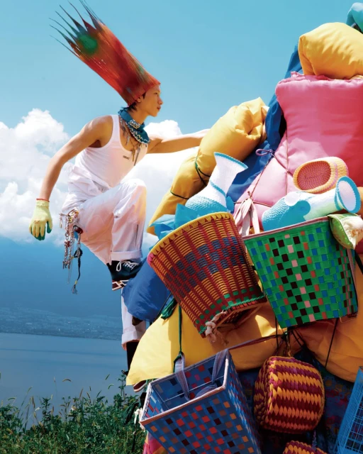 Person in white outfit climbing a colorful tower of pillows and woven baskets against a bright sky and distant view of water.