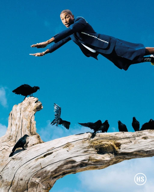Pharrell Williams in mid-air wearing a navy suit with crows perched on a weathered branch against a bright blue sky. Fashion editorial.