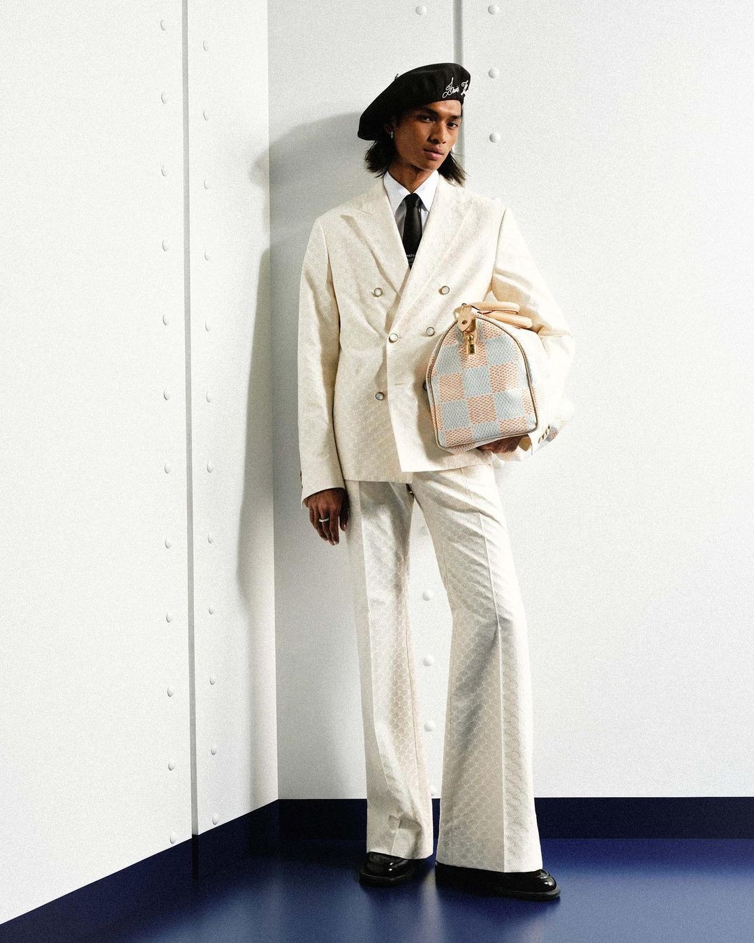 Man poses in cream-colored suit with patterned bag indoors. The man wears a black beret, and is on a blue and white surface.