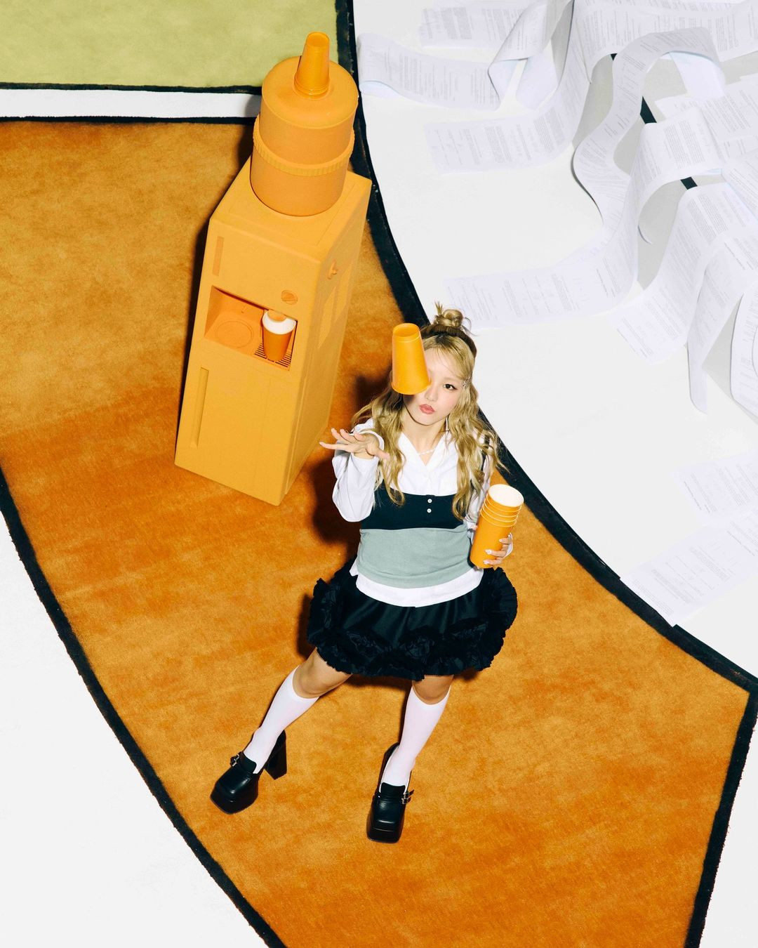 Young woman in playful studio shot balances orange cups while standing on a carpet next to a toy refrigerator, styled with white tights and chunky shoes.