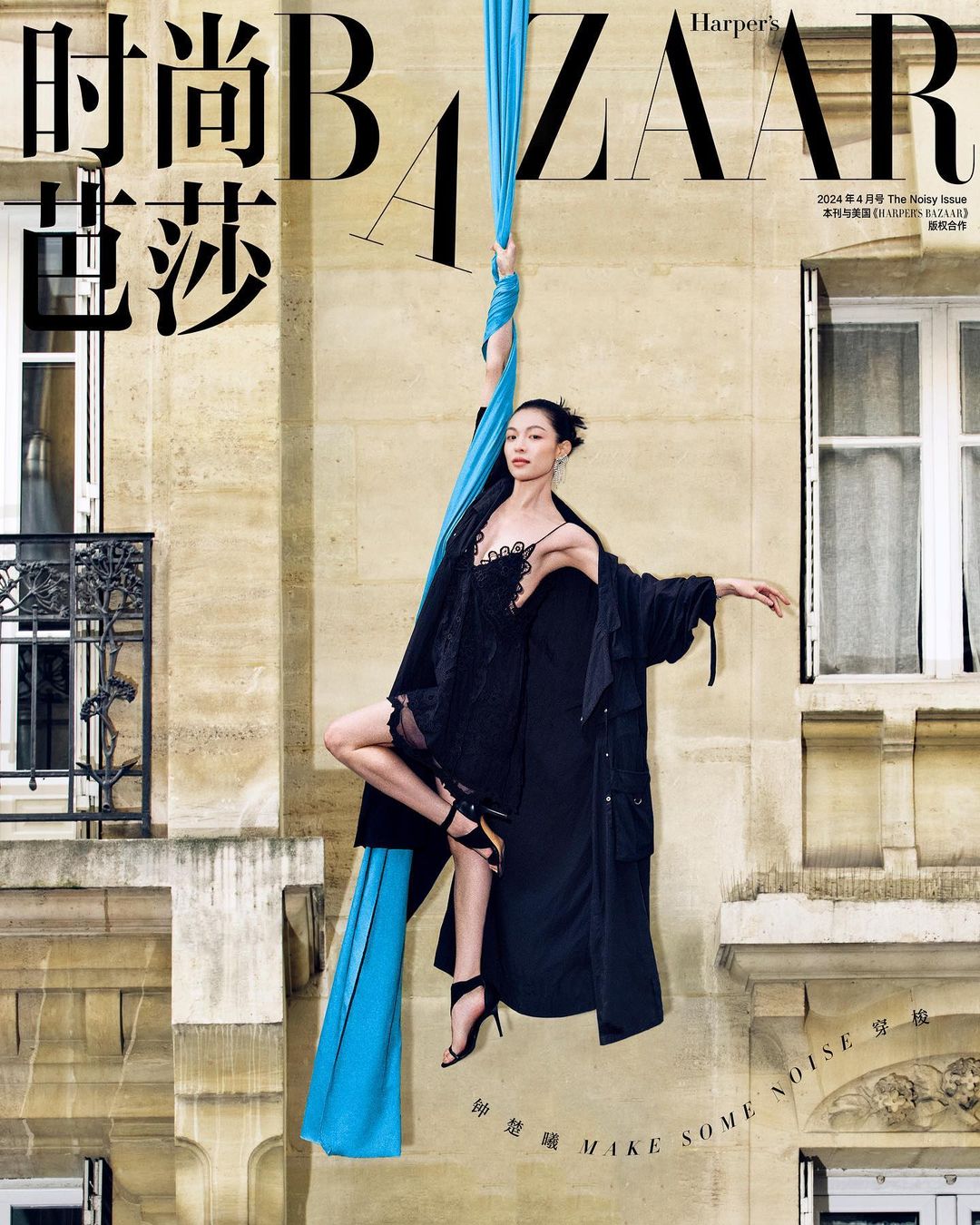 Fashion editorial cover featuring a woman suspended in air, wearing a black lace dress and long jacket, holding onto a blue aerial silk.
