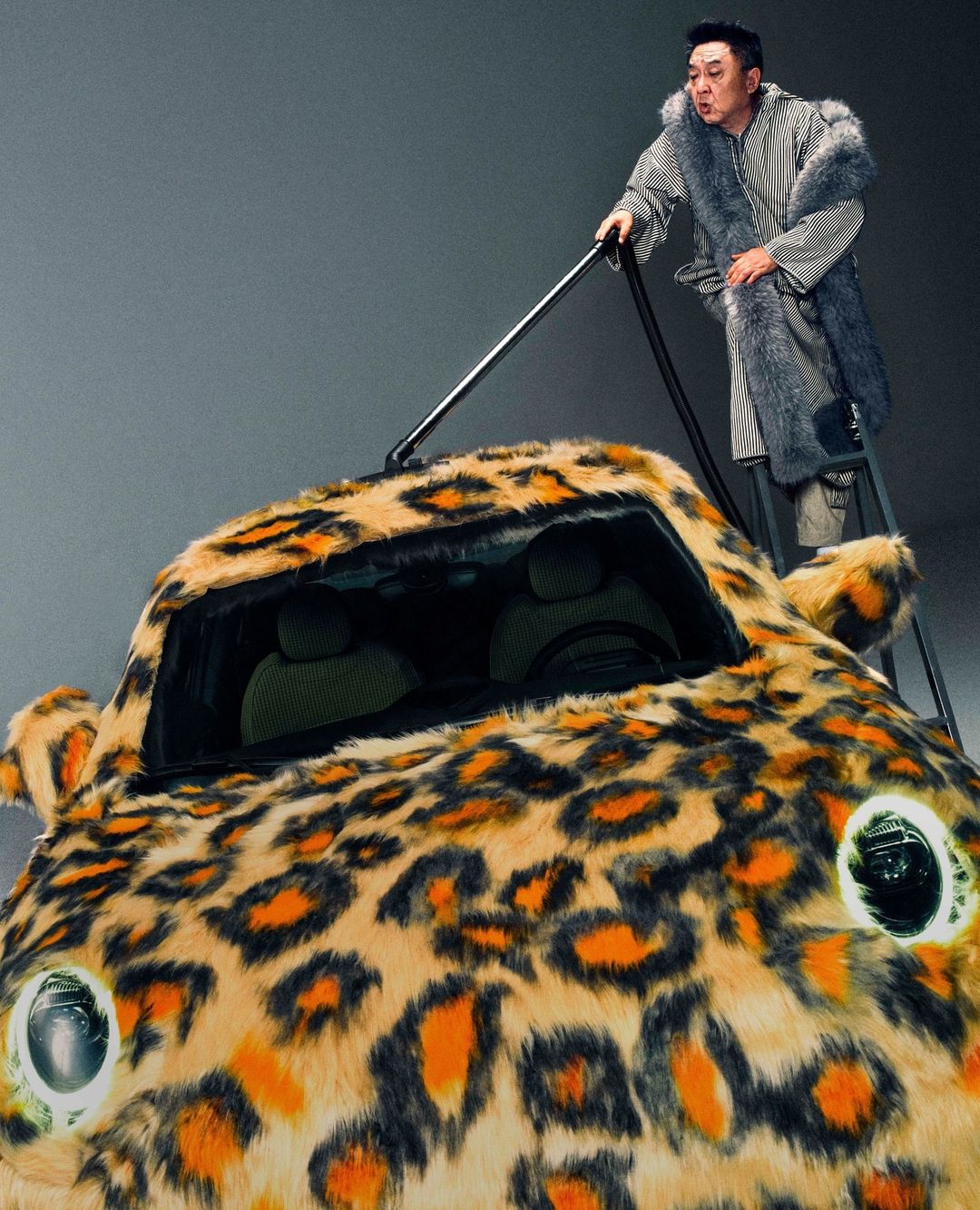 A man standing on a ladder vacuums a small car covered in a fur-like, leopard-print fabric in a grey, indoor studio setting.