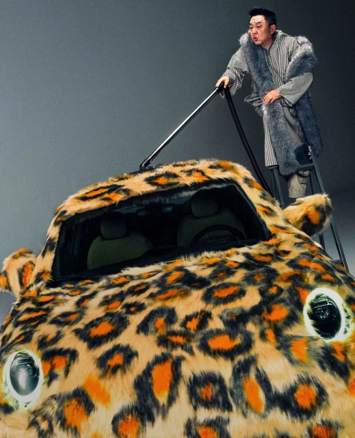 A man standing on a ladder vacuums a small car covered in a fur-like, leopard-print fabric in a grey, indoor studio setting.
