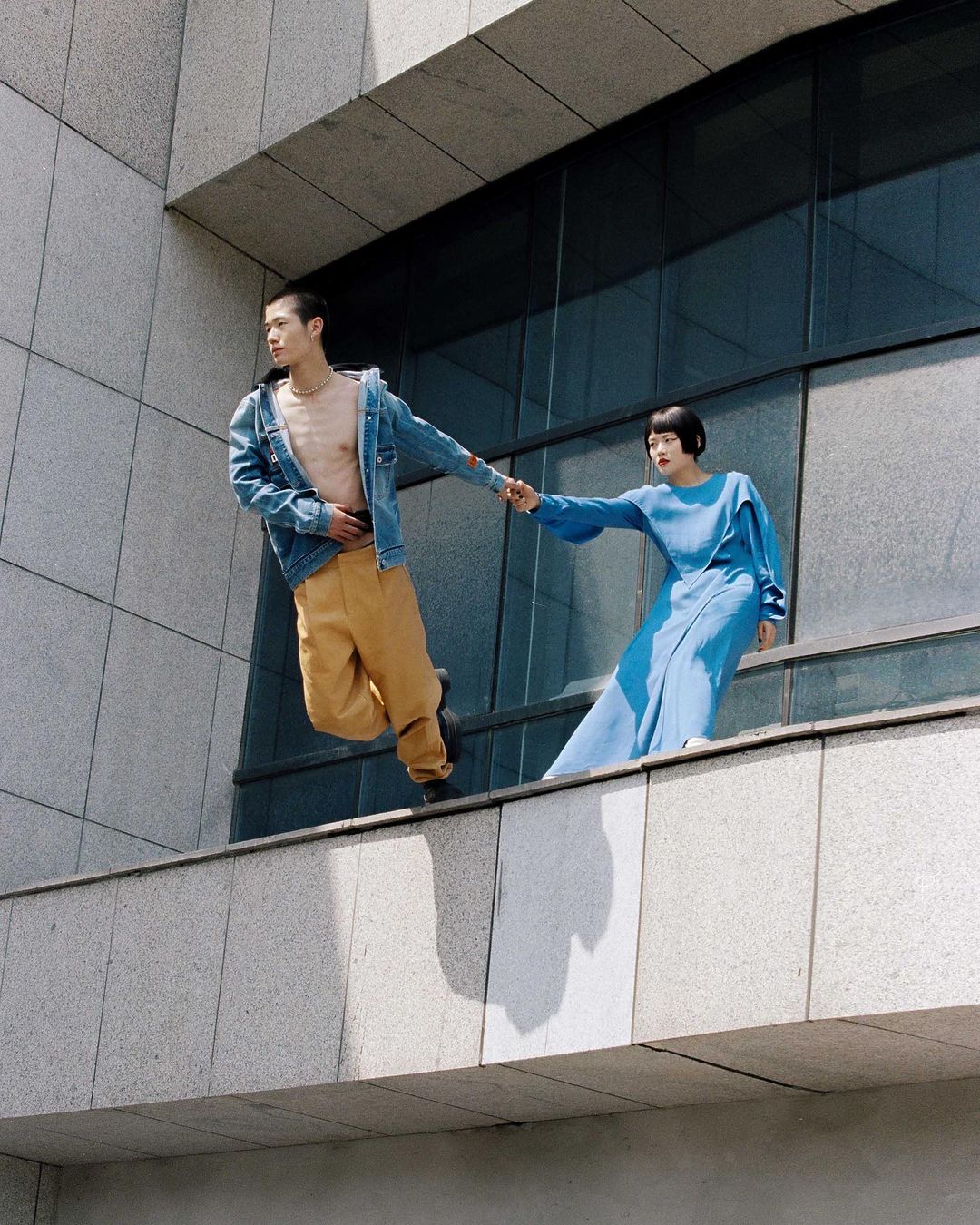 A stylish couple poses on a building ledge, the man in a denim jacket and the woman in a blue dress, set against a modern, architectural backdrop.