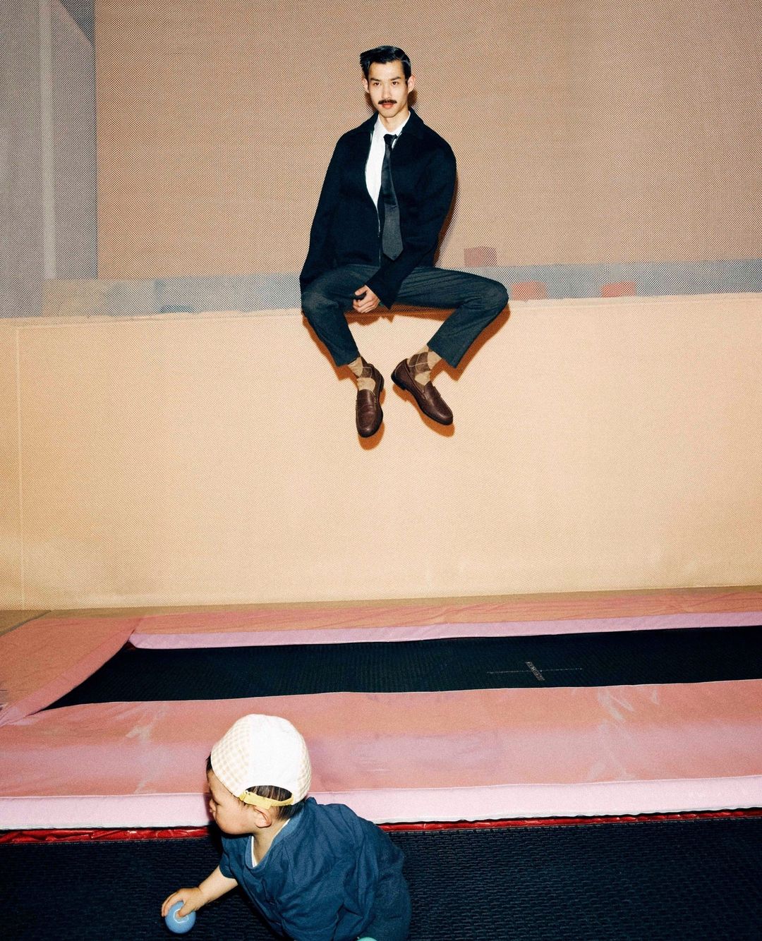 A man in a blazer and tie sits high on a ledge over a trampoline and child, creating a playful juxtaposition of formal wear and recreation.