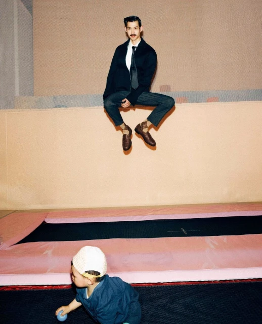 A man in a blazer and tie sits high on a ledge over a trampoline and child, creating a playful juxtaposition of formal wear and recreation.