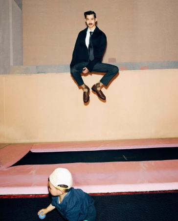 A man in a blazer and tie sits high on a ledge over a trampoline and child, creating a playful juxtaposition of formal wear and recreation.