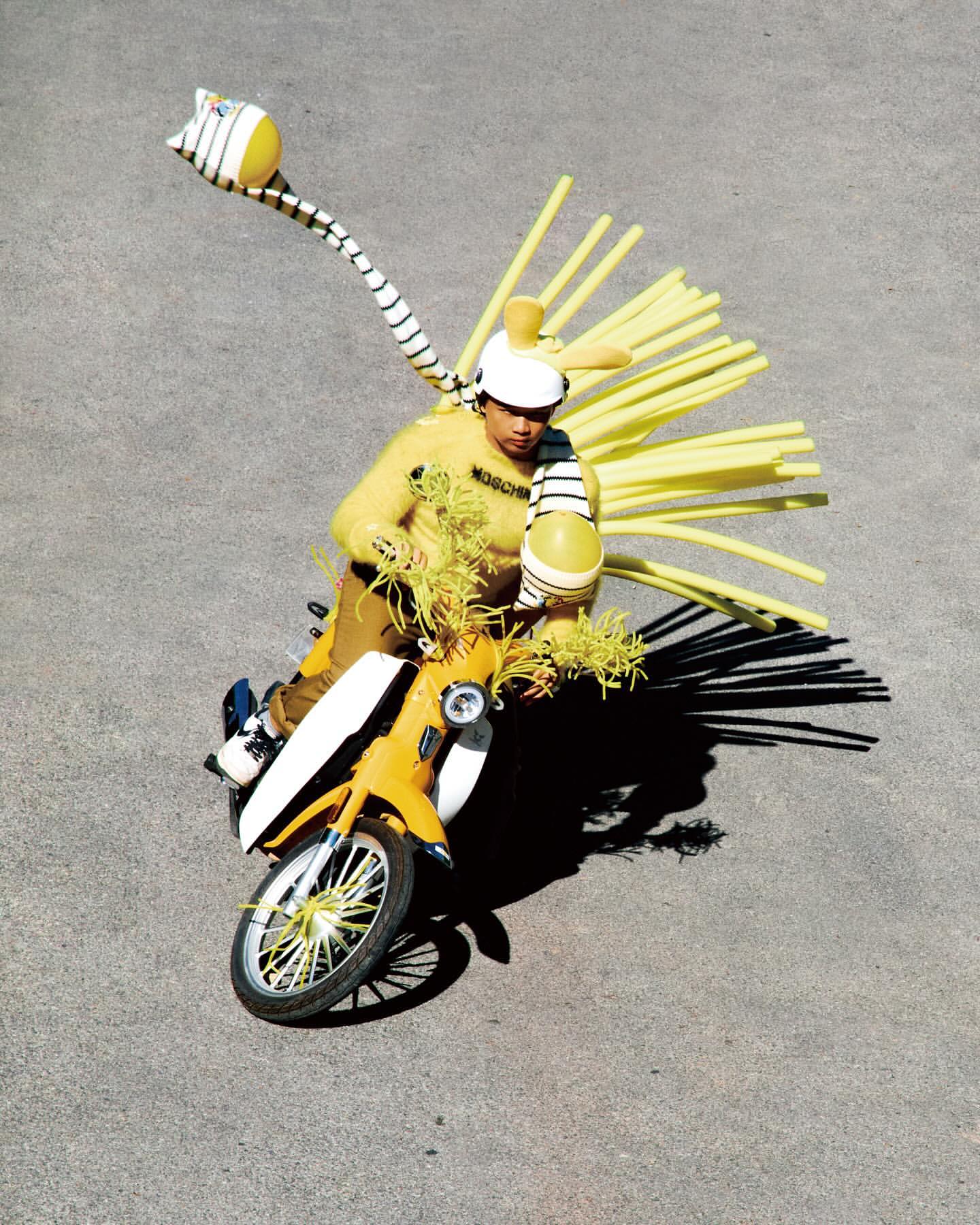 A person in a yellow sweater and a white helmet with yellow tubes on a yellow moped on a concrete surface in an outdoor fashion shoot.