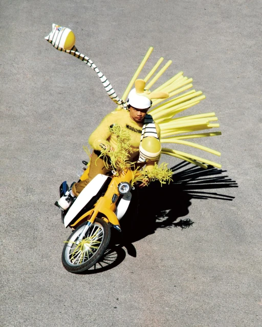 A person in a yellow sweater and a white helmet with yellow tubes on a yellow moped on a concrete surface in an outdoor fashion shoot.