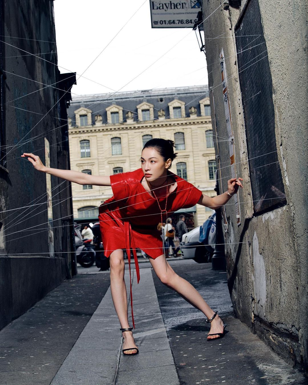 A woman in a red dress poses in an urban alleyway, framed by hanging strings, featuring a building in the background.
