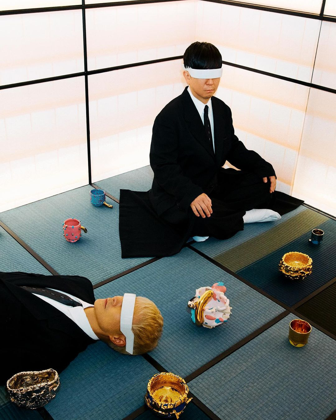Two Asian people in suits with white blindfolds are on a tatami mat in front of a wall panel with geometric black accents.