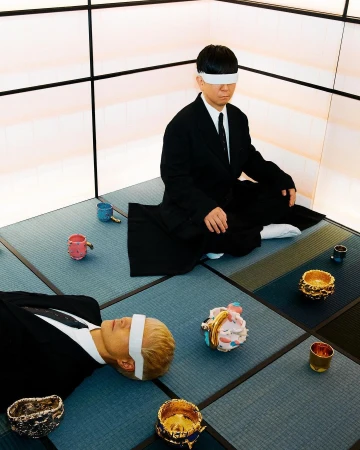 Two Asian people in suits with white blindfolds are on a tatami mat in front of a wall panel with geometric black accents.