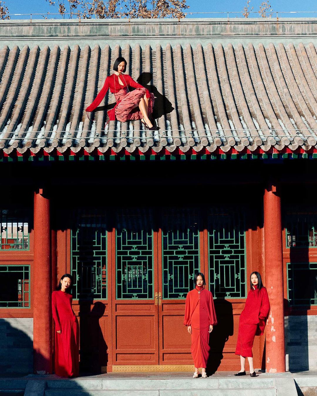 Four Asian women in red dresses pose in front of a traditional Chinese building with a tiled roof under a clear blue sky, fashion photoshoot.