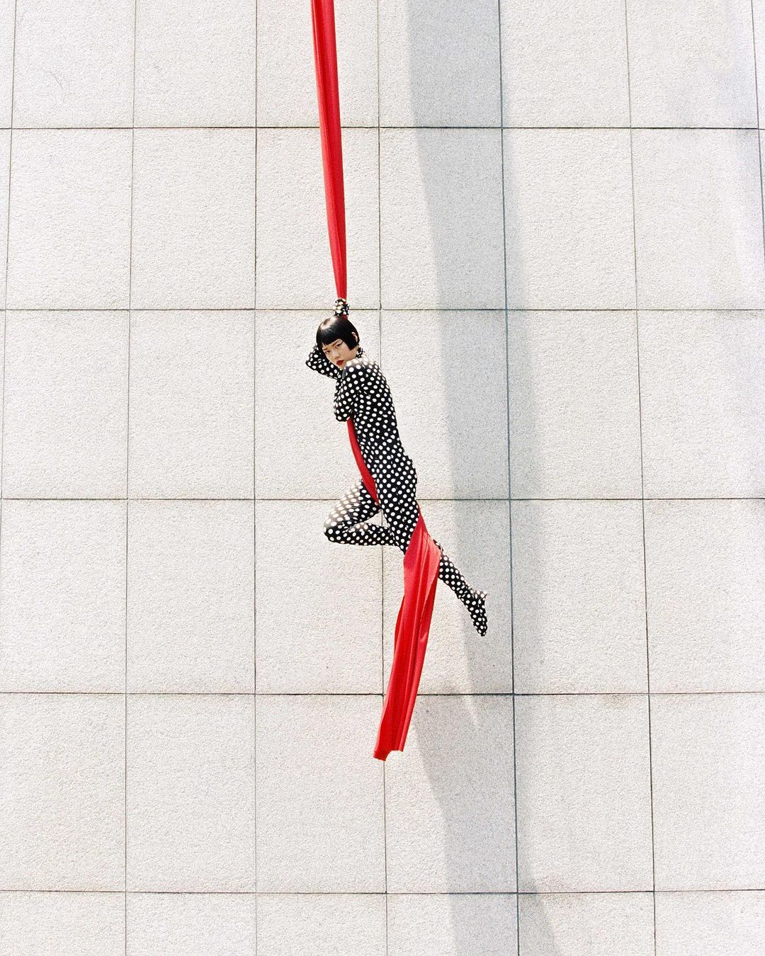 A woman in a polka dot bodysuit performs aerial silks on a red silk ribbon against a textured grey wall, captured in an eye-level medium shot.