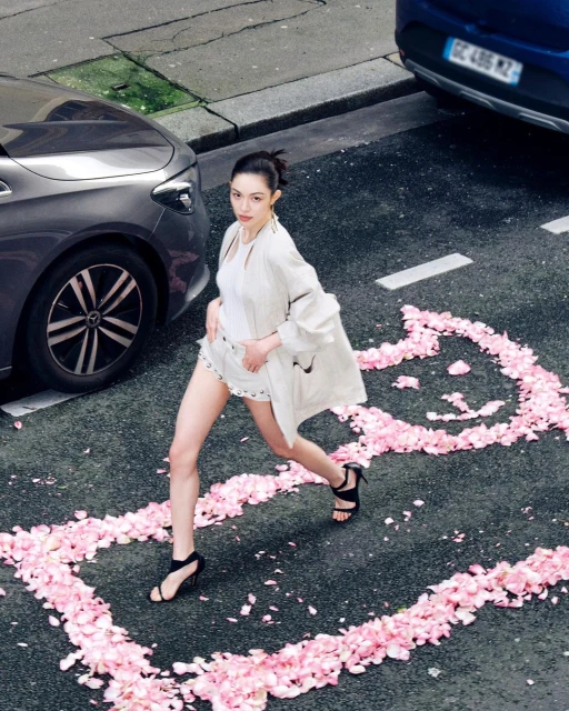 Adult female fashion model in neutral clothing and heels walks on asphalt with cars, surrounded by a border of pink flower petals.