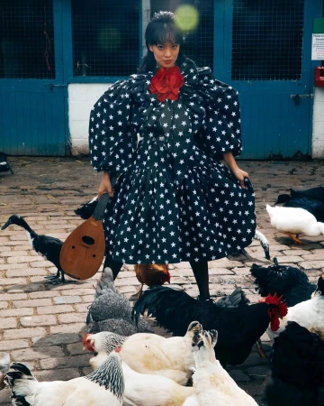 Woman in a star-patterned dress standing amidst chickens in a brick-paved farmyard, holding a stringed instrument with a barn door behind.
