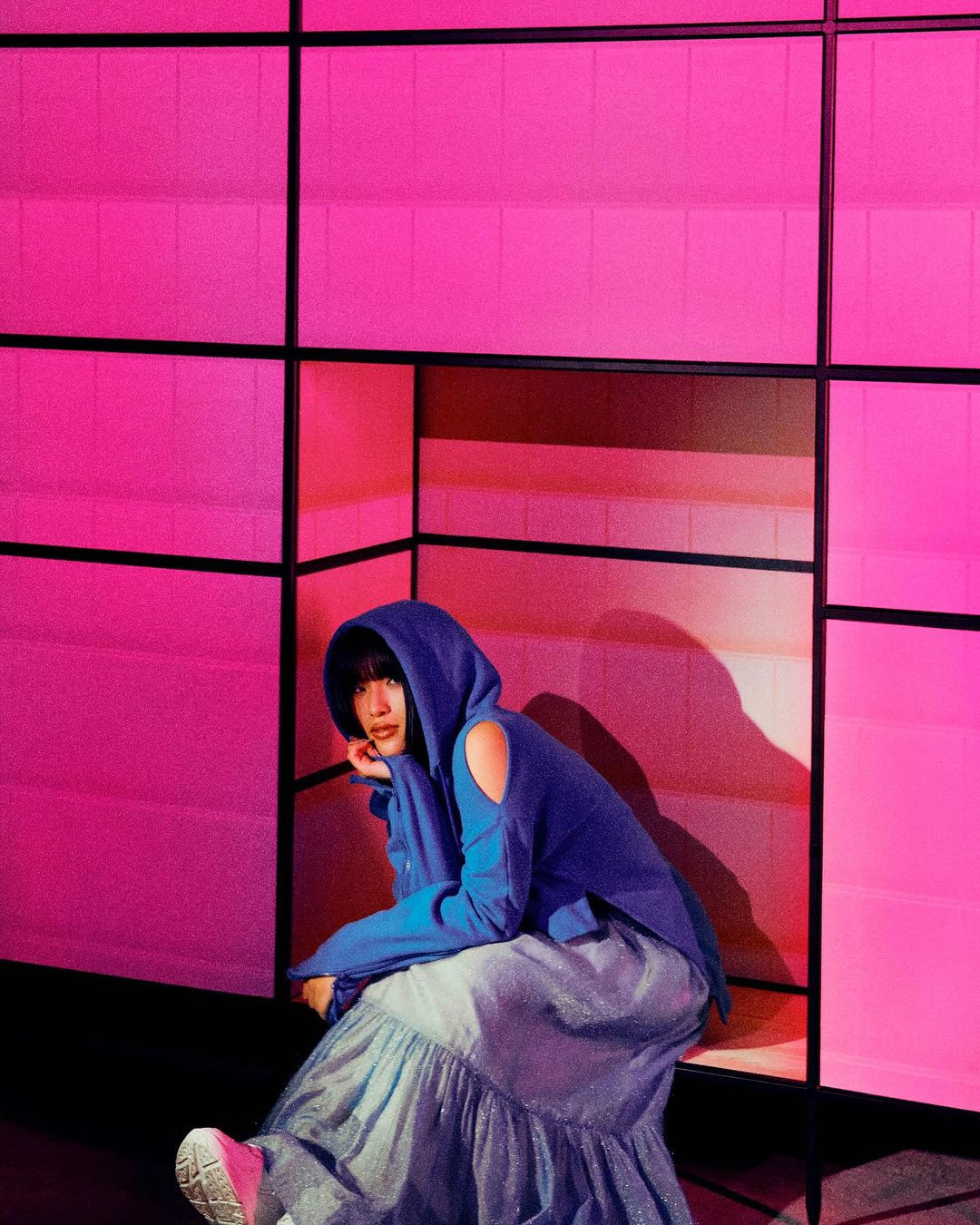 Young adult woman with bangs wearing a blue hooded sweatshirt and skirt sitting in front of a modern, pink geometric background.