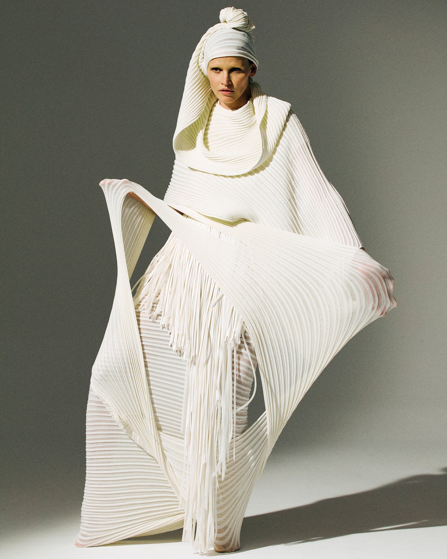 Woman in an all-white pleated gown with head wrap posing in studio, dramatic draping and monochrome aesthetic for fashion editorial.