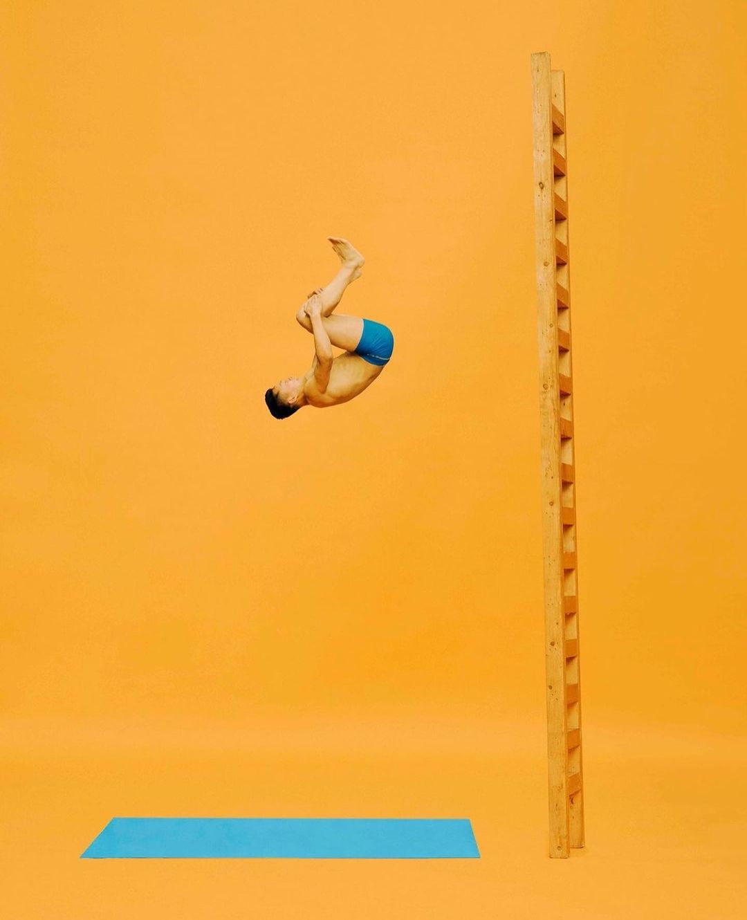 Young man in mid-air performing acrobatics in front of a ladder with a light blue mat on an orange seamless background.