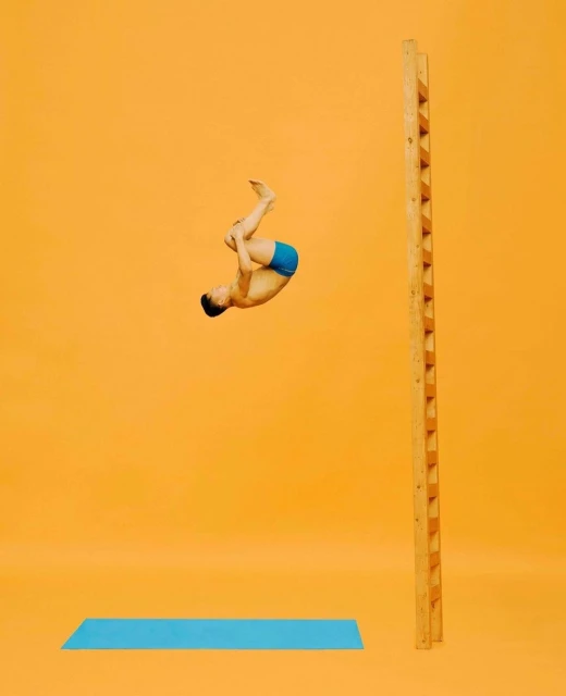 Young man in mid-air performing acrobatics in front of a ladder with a light blue mat on an orange seamless background.