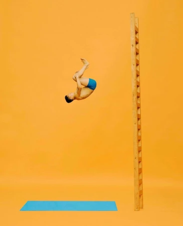 Young man in mid-air performing acrobatics in front of a ladder with a light blue mat on an orange seamless background.