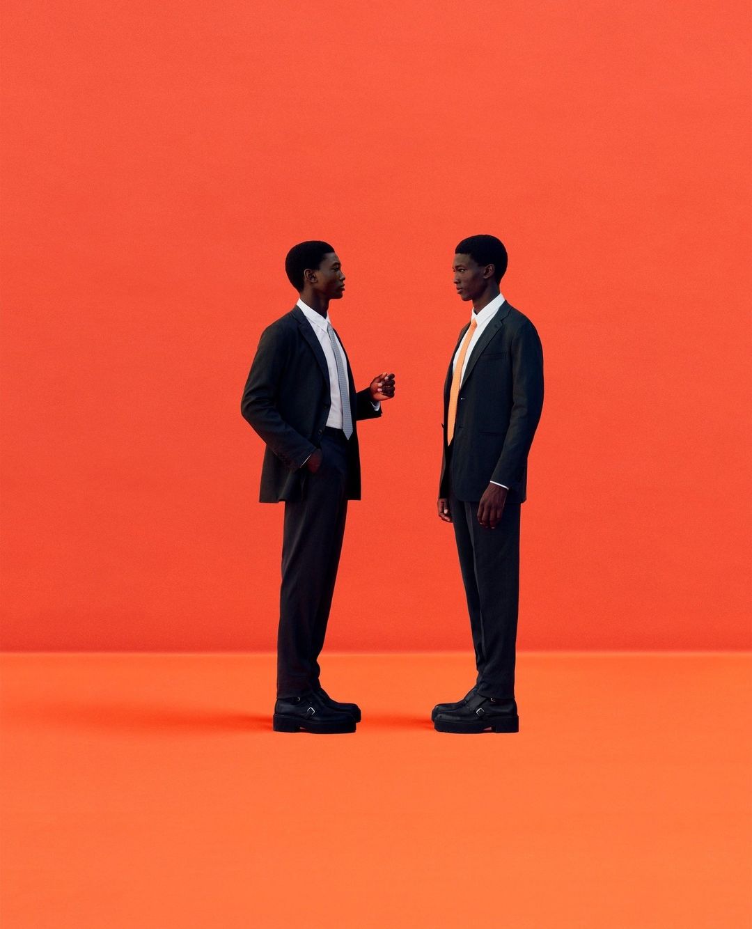 Two Black men in suits stand facing each other against an orange backdrop. Minimalist composition, studio lighting, professional attire, and dark shoes.