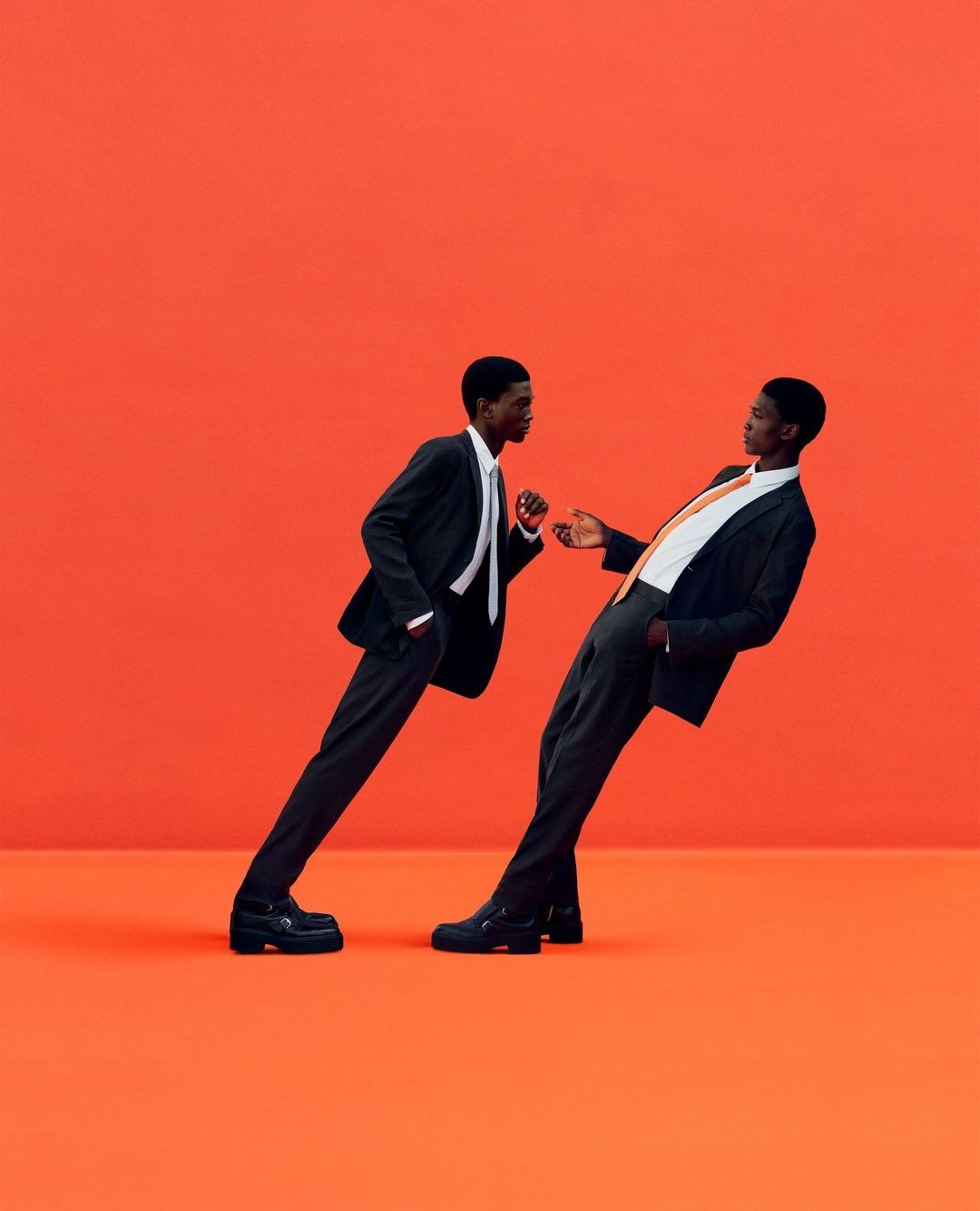 Two African American men in dark suits lean in toward each other on an orange background, creating a striking and modern image.