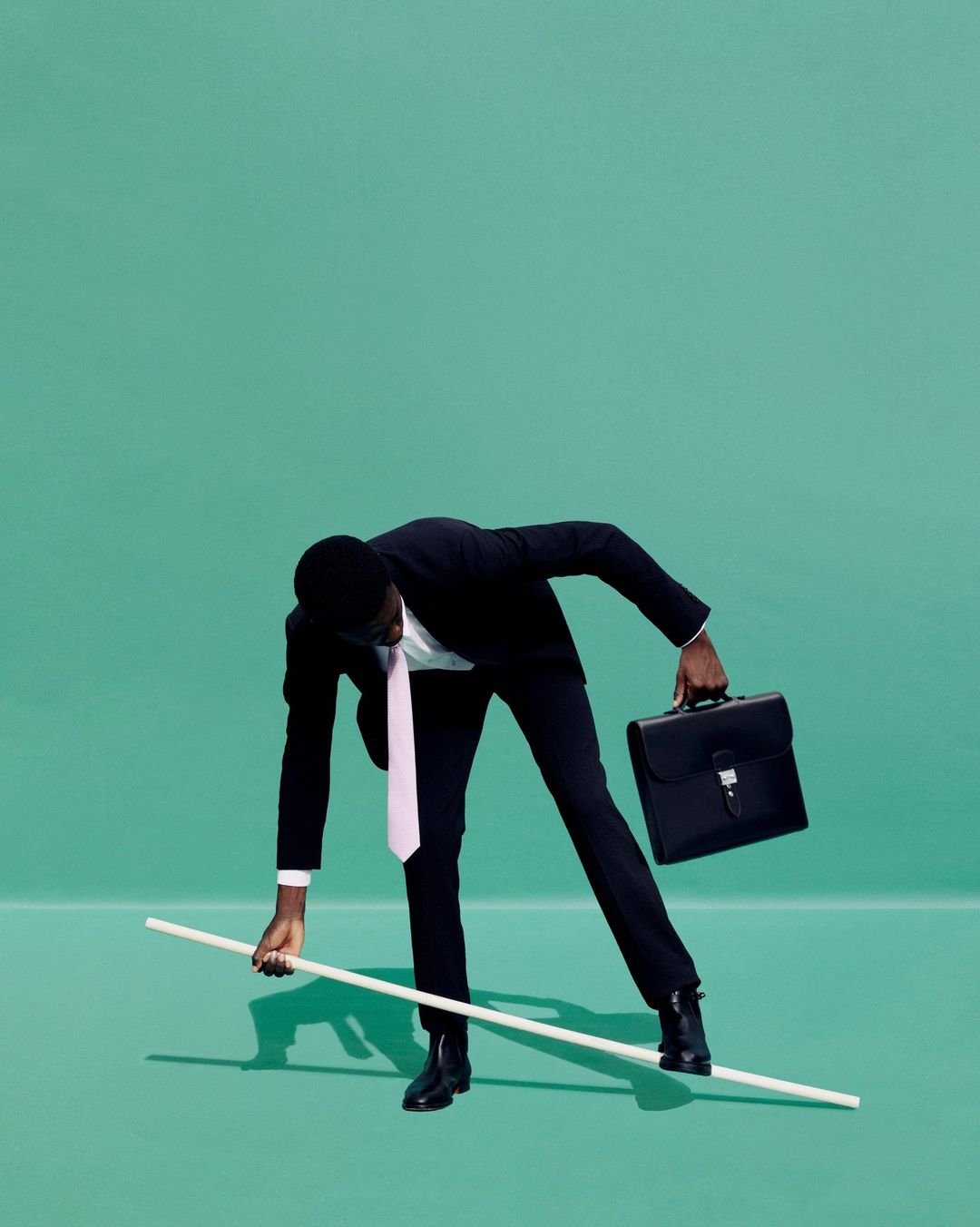 An African-American man in a suit balances on a white stick, holding a briefcase, against a solid green backdrop in a studio setting.