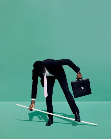 An African-American man in a suit balances on a white stick, holding a briefcase, against a solid green backdrop in a studio setting.