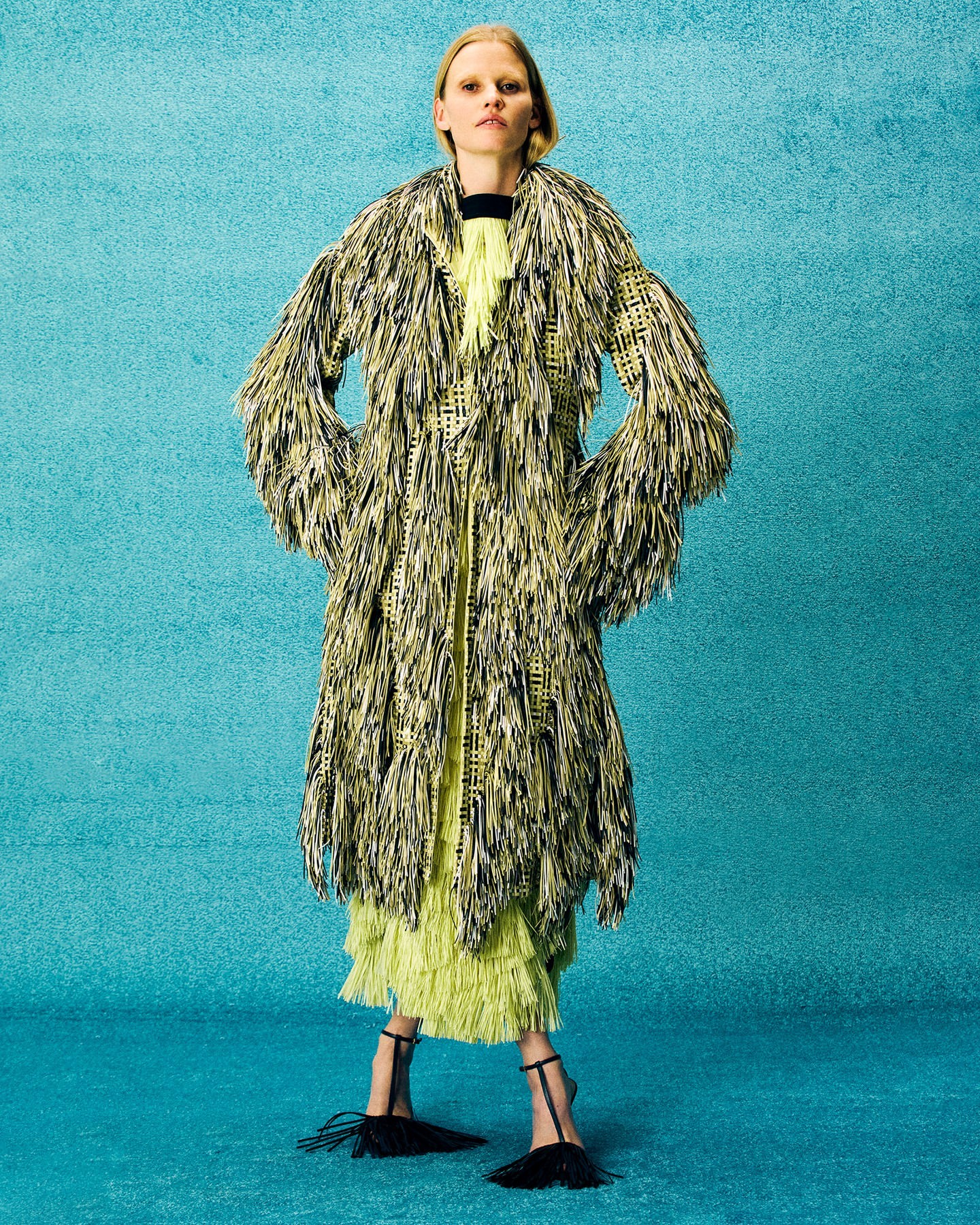 Fashionable adult female model poses in full shot, wearing an avant-garde coat with fringe, a dress, and heeled shoes against a teal background.