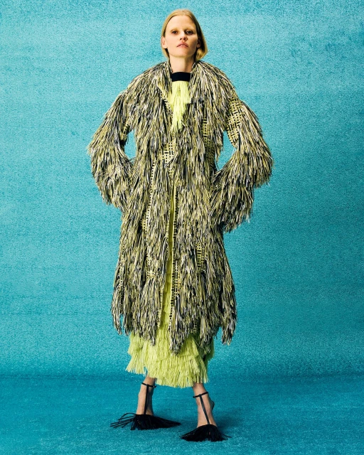Fashionable adult female model poses in full shot, wearing an avant-garde coat with fringe, a dress, and heeled shoes against a teal background.
