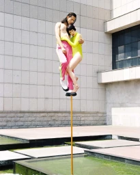 Man and woman balanced precariously atop a tall thin pole above water, photographed by Julian Song.