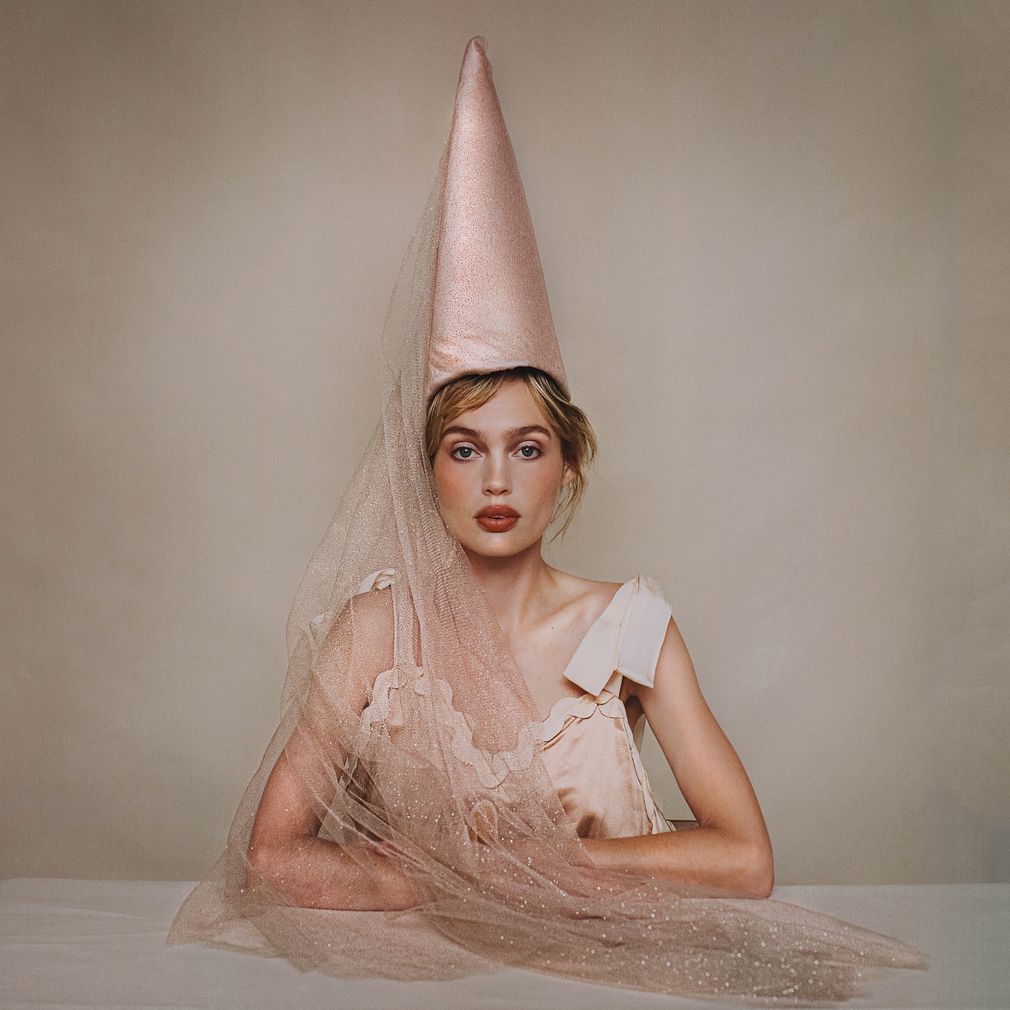 Young woman wearing a cone-shaped hat with a veil poses indoors against a neutral backdrop, soft lighting enhances her features.