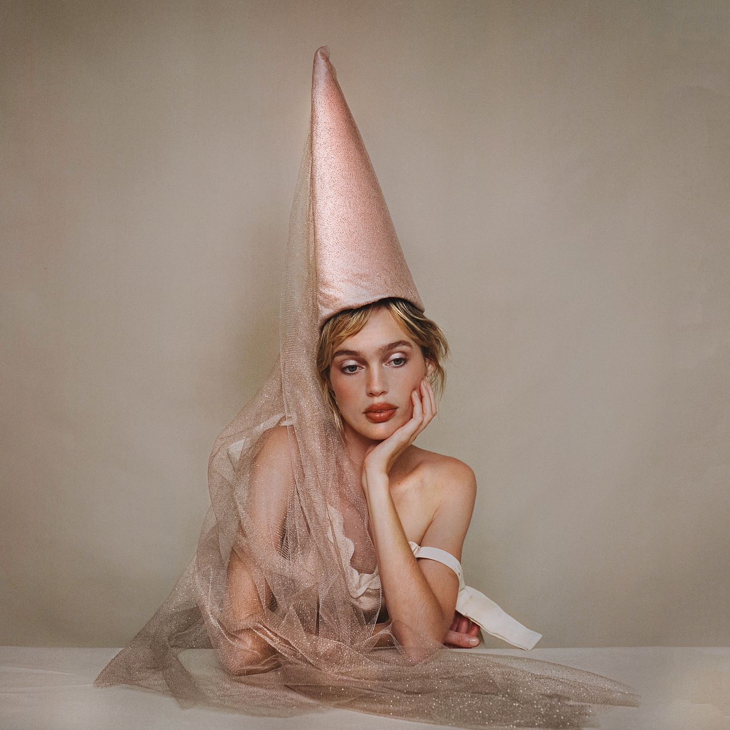 Elegant woman poses in a stylish cone-shaped hat with sheer fabric against a soft neutral backdrop, emphasizing beauty and artistic fashion.