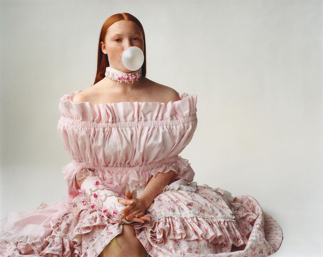 Red-haired woman in an elaborate pink off-shoulder dress blows a bubble with bubblegum in a photo studio setting with a white background.