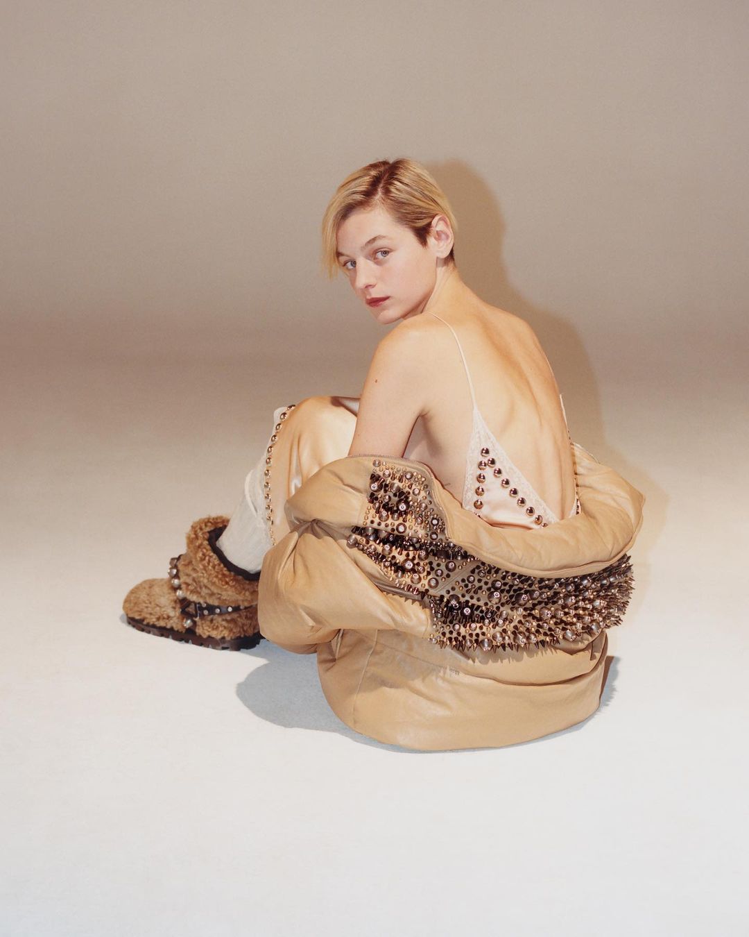 Portrait of a woman with short blonde hair wearing a beige dress and oversized jacket in a studio setting looking over shoulder.