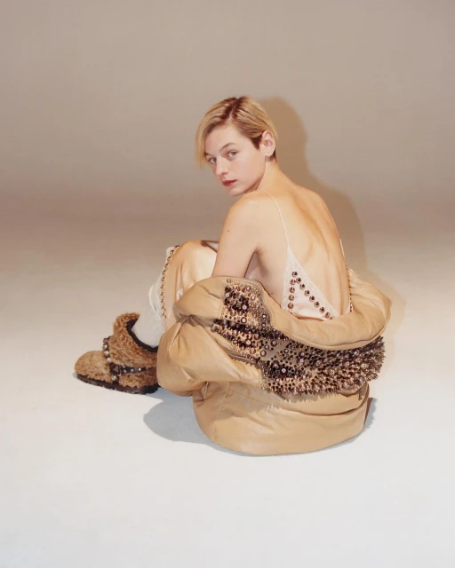Portrait of a woman with short blonde hair wearing a beige dress and oversized jacket in a studio setting looking over shoulder.