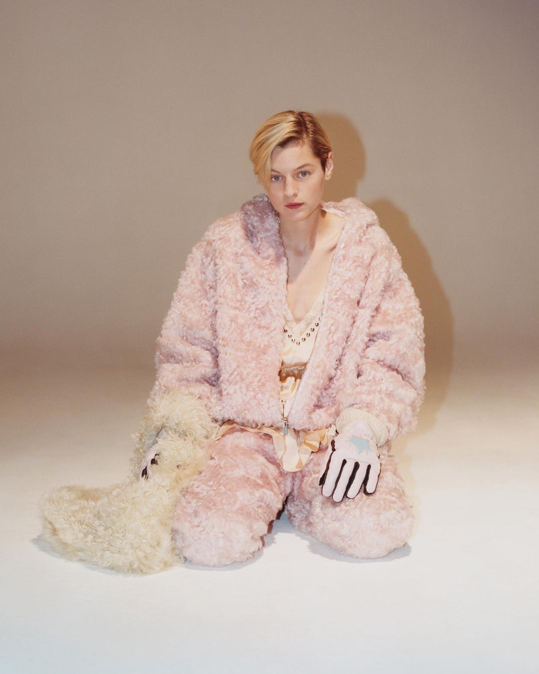 Woman in a pink fuzzy jacket and pants sits in a photo studio with a serious facial expression, wearing white gloves and white boots.