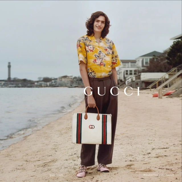 A fashionable young man in a patterned shirt and brown slacks holds a Gucci tote bag on a sandy beach near a coastal town.