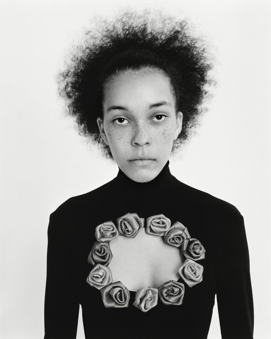 Portrait of a woman with afro hair wearing a black turtleneck sweater with rose embellishments, set against a stark white background.