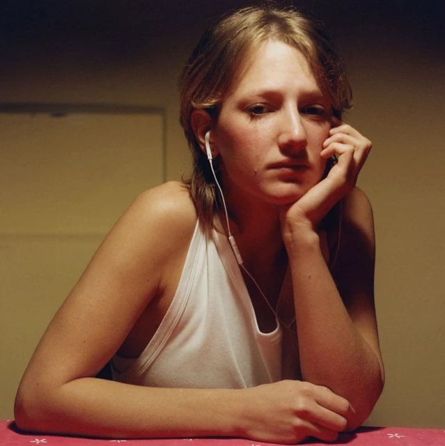 Blonde woman in a white tank top wearing earphones, resting her head on her hand while sitting at a table indoors, looking downward.