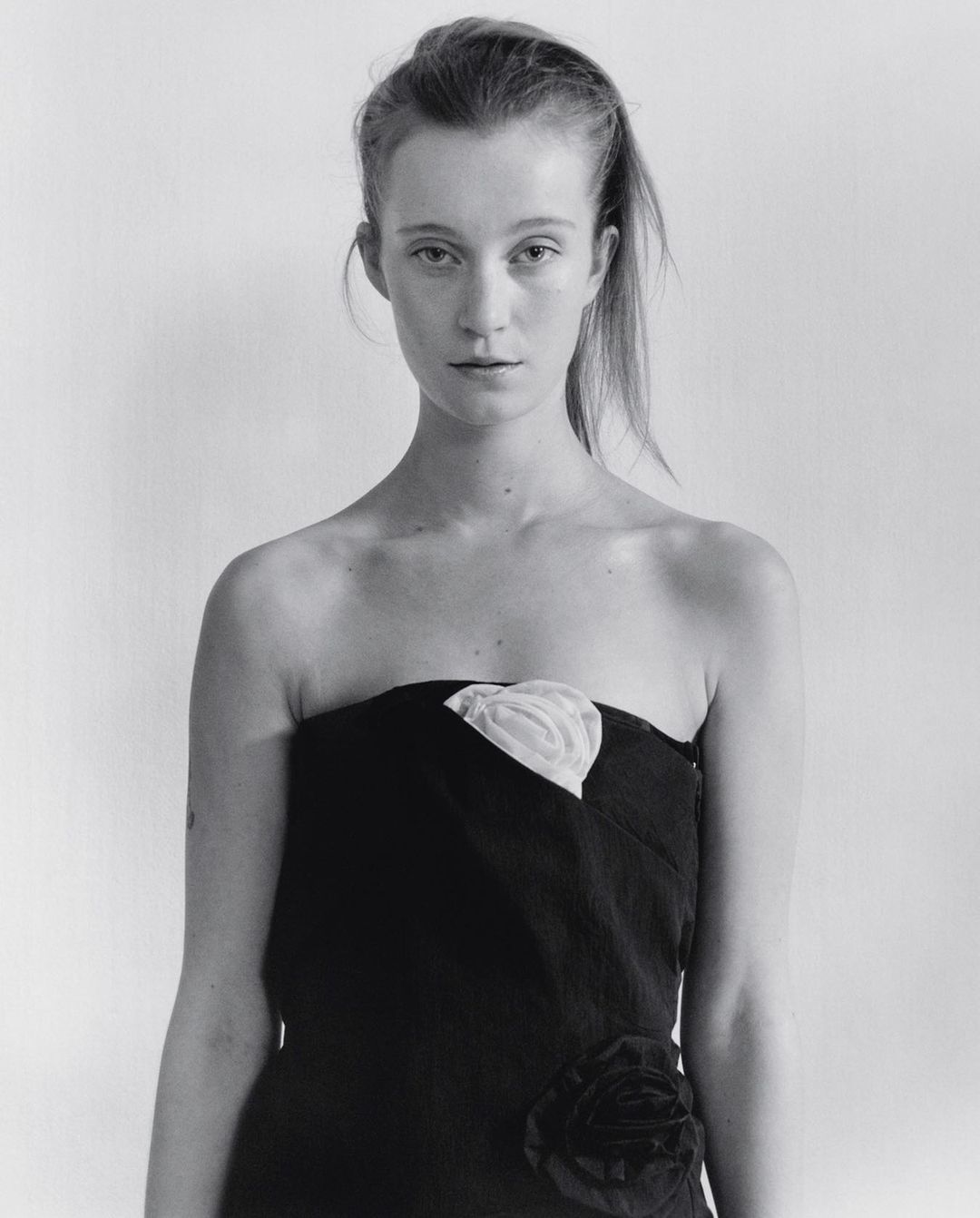 Monochrome portrait of a woman with long hair, wearing a strapless black dress with a white floral detail, against a white background.